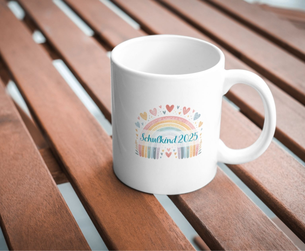 Weiße Keramiktasse mit pastellfarbenem Regenbogen, Herzchen und dem Schriftzug „Schulkind 2025“ in Hellblau, fotografiert auf einem Holztisch.